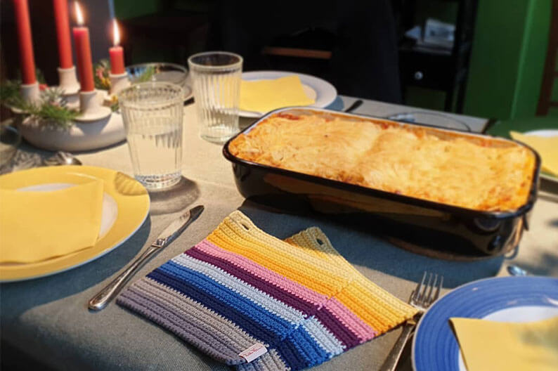 Bunte gehäkelte Topflappen liegen auf einem gedeckten Esstisch neben einer Auflaufform.