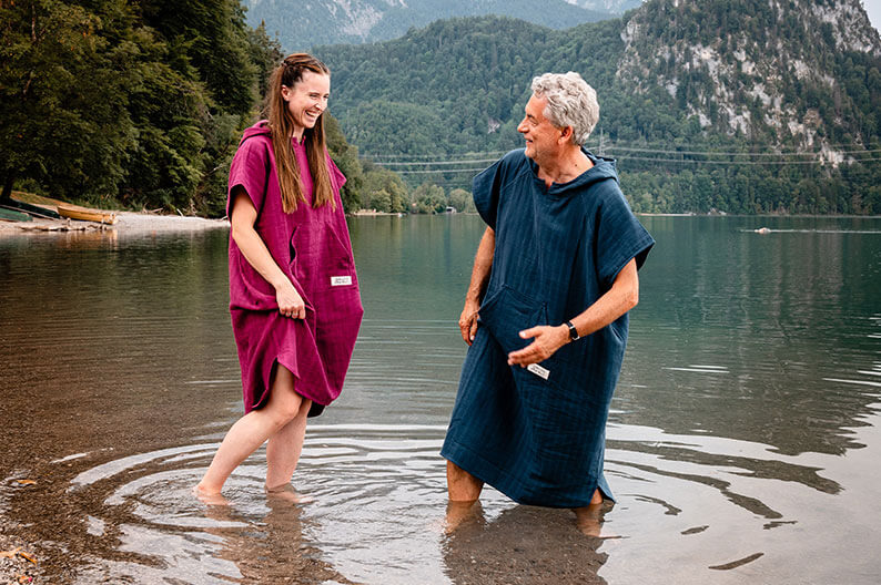 Zwei Menschen in pinkfarbenem und dunkelblauem Strandponcho stehen lachend im flachen Wasser eines Bergsees.