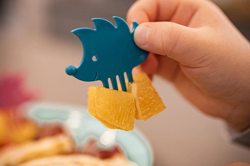Eine Kinderhand hält einen kleinen blauen Snack-Picker in Igelform mit einem aufgespießten Stück Obst.