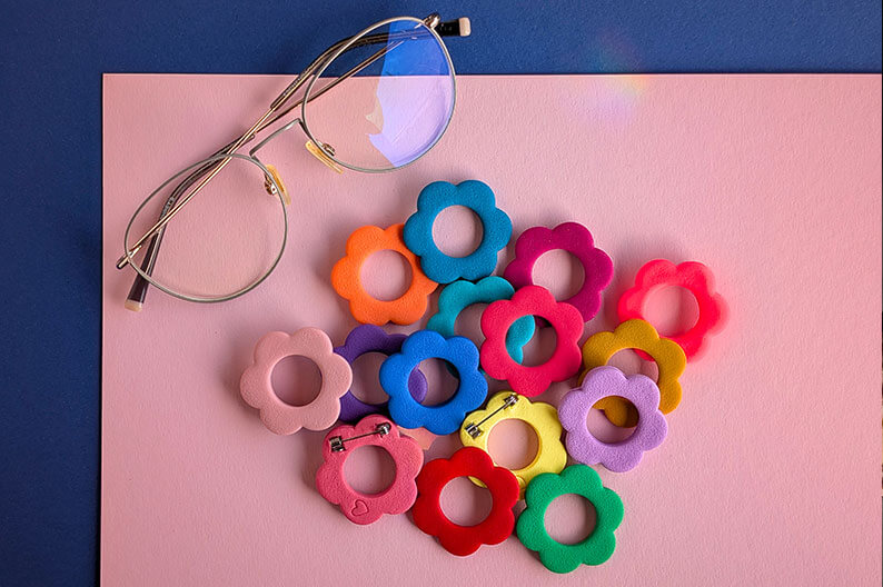 Bunte Brillenbroschen in Blumenform liegen zusammen mit einer Brille auf einem zweifarbigen Hintergrund.