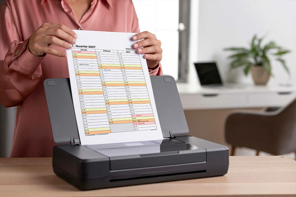 Eine Person hält einen ausgedruckten Quartalskalender 2027 von Kalenderpedia an einem Drucker