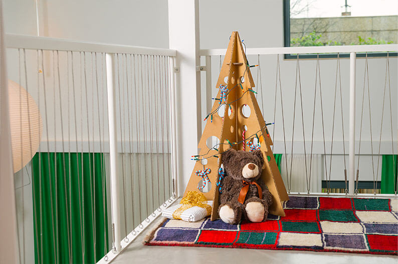 Geschmückter Weihnachtsbaum aus Pappe steht in einem hellen Raum auf einer Galerie.