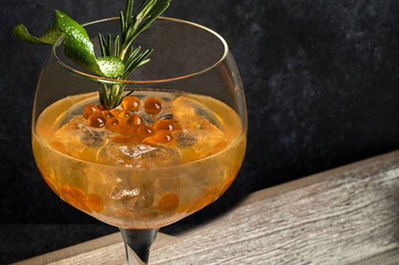 Cocktail in einem Glas mit orangefarbenen Gourmet-Perlen und Rosmarinzweig vor dunklem Hintergrund.