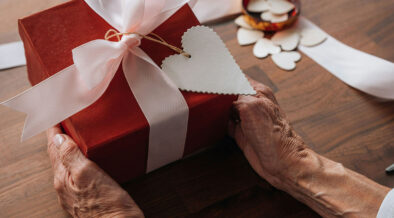 Ältere Hände halten ein rot verpacktes Geschenk mit weißer Schleife und herzförmigem Anhänger.