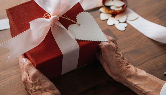 Ältere Hände halten ein rot verpacktes Geschenk mit weißer Schleife und herzförmigem Anhänger.