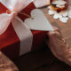 Ältere Hände halten ein rot verpacktes Geschenk mit weißer Schleife und herzförmigem Anhänger.
